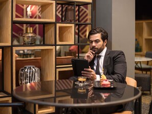 Een man in pak zit aan een ronde tafel met een laptop, een drankje en een bord eten. Op de achtergrond een houten boekenkast en een modern interieur.