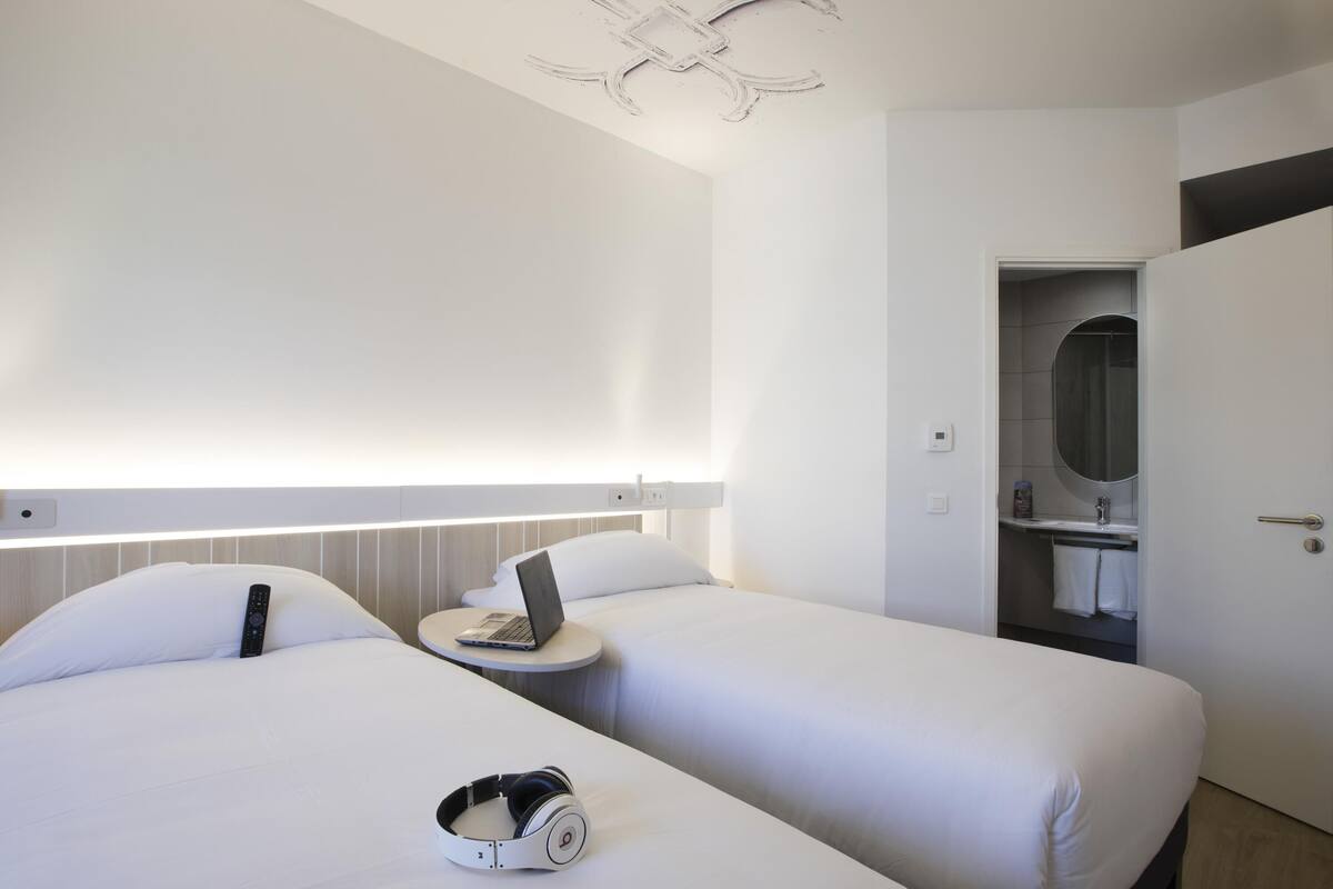 Een hotelkamer met twee bedden, een laptop op een nachtkastje, een koptelefoon, een ronde spiegel en een deur naar een badkamer.