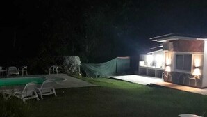 A nighttime view of a backyard with a swimming pool, a covered area, and a small building with lights.