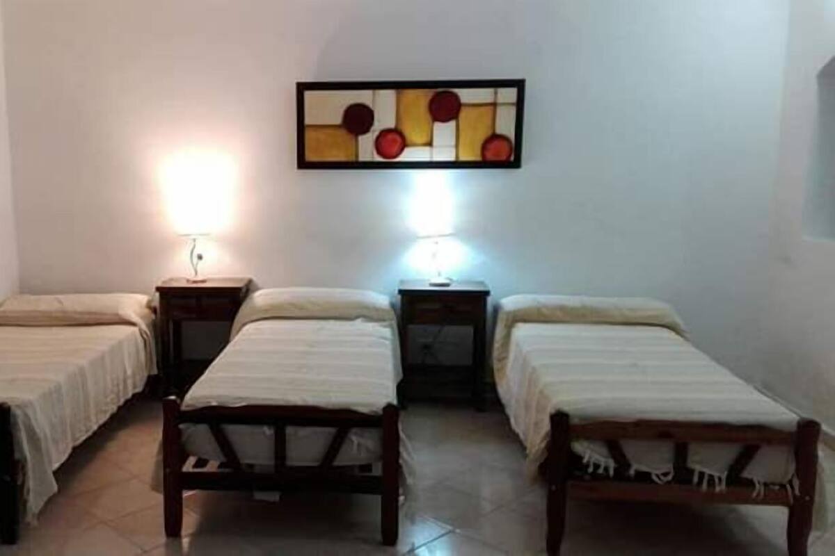 Une chambre avec trois lits simples, chacun doté d’un cadre en bois et de draps blancs. On y trouve deux tables de chevet avec des lampes, et une œuvre d’art abstraite accrochée au mur au-dessus des lits.