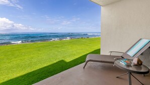 A balcony with a view of the ocean, a lounge chair, and a small table with books and a shell. - Oceanfront Two Bedroom Condo (Koloa)