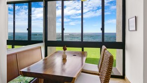 A room with a large window overlooking the sea, a wooden table, and a woven chair. - Oceanfront Two Bedroom Condo (Koloa)