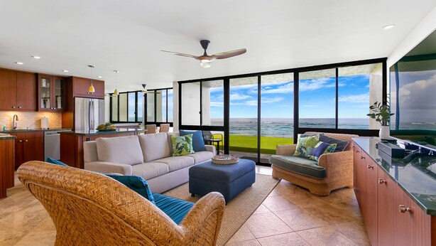 A spacious living area with a large window overlooking a scenic view, a kitchen with modern appliances, and comfortable seating. - Oceanfront Two Bedroom Condo (Koloa)