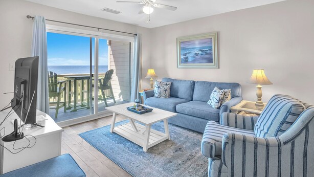 Een woonkamer met een bank, fauteuil, salontafel en een televisie. Er is een balkon met uitzicht op de oceaan.
