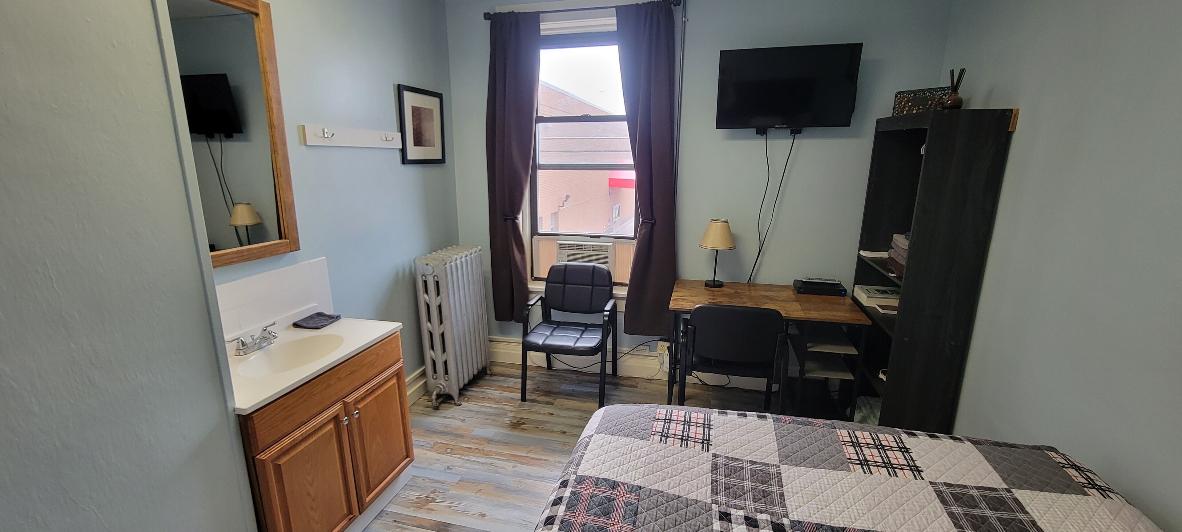 A small, well-lit room with a bed, a desk, a chair, a radiator, a TV, and a window with curtains.