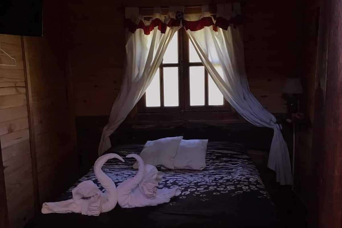 A wooden room with a bed, white curtains, and a window.