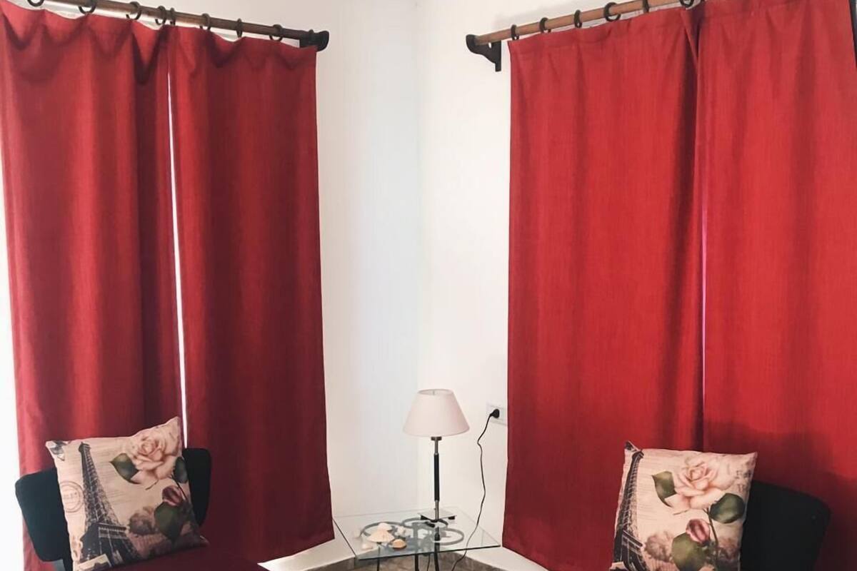 A room with two armchairs, a small table with a lamp, and red curtains.