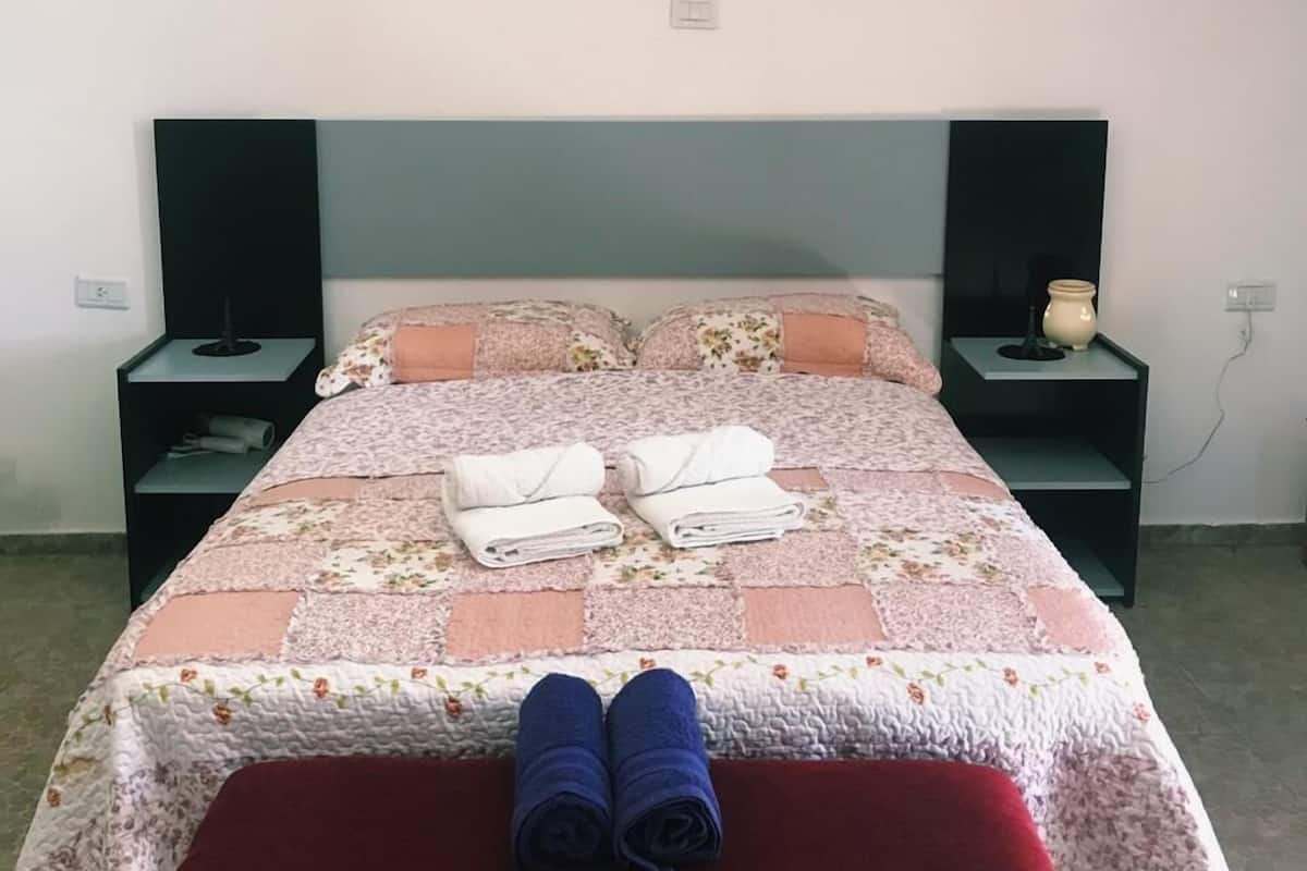 A neatly made bed with a floral bedspread, two rolled towels, and a red bench in front. There are two bedside tables with lamps and a power outlet on the wall.