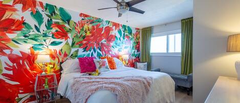 A hotel room with a large bed, a ceiling fan, a window with curtains, and a colorful wall mural.