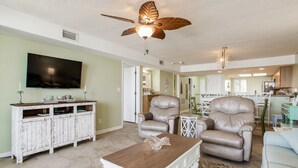 A spacious living room with a ceiling fan, a flat-screen TV, leather armchairs, a coffee table, and a dining area with a kitchen in the background. - Orange Beach Gulf-Front 2BR • Sleeps 7 • 2 La-Z-Boy Recliners • Pools • Boats (Orange Beach)
