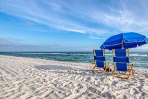Two blue beach chairs and a blue umbrella on a sandy beach with the ocean in the background.
