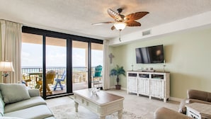 A spacious living room with a large sliding glass door leading to a balcony, a ceiling fan, a flat-screen TV mounted on the wall, and a white wooden cabinet. - Orange Beach Gulf-Front 2BR • Sleeps 7 • 2 La-Z-Boy Recliners • Pools • Boats (Orange Beach)