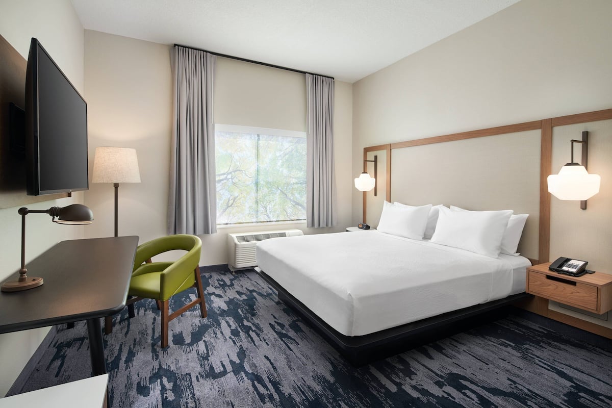 A hotel room with a bed, a desk, a chair, a TV, a lamp, a nightstand, and a window with curtains.