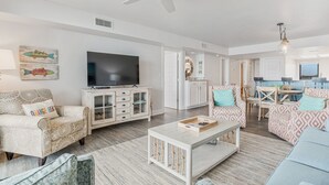A spacious living room with a ceiling fan, a TV, a dining area with a table and chairs, and a large window. - Orange Beach Gulf View 2BR • Sleeps 6 • 2 Pools • Beach • Boat Slips (Orange Beach)