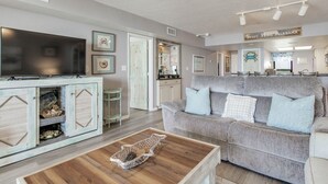 Un salon avec un canapé gris, une table basse en bois et un téléviseur posé sur un meuble. On aperçoit, au fond, un coin cuisine avec une enseigne sur laquelle on peut lire : « Sweet Home Alabama ».