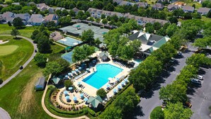 Vista aérea de una zona residencial con una gran piscina rodeada de sillones de descanso y sombrillas, una cancha de tenis y un club social.