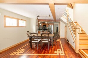 Un comedor amplio con una mesa y sillas de madera, una cocina al fondo y una escalera de madera.