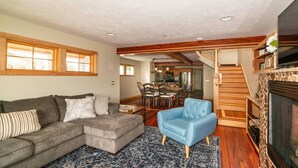 A living room with a grey sectional sofa, a blue armchair, and a patterned rug. There is a wooden staircase and a fireplace with a stone surround. - Second Street Stay–Downtown+ WALKABLE TO CHERRY FEST (Traverse City)
