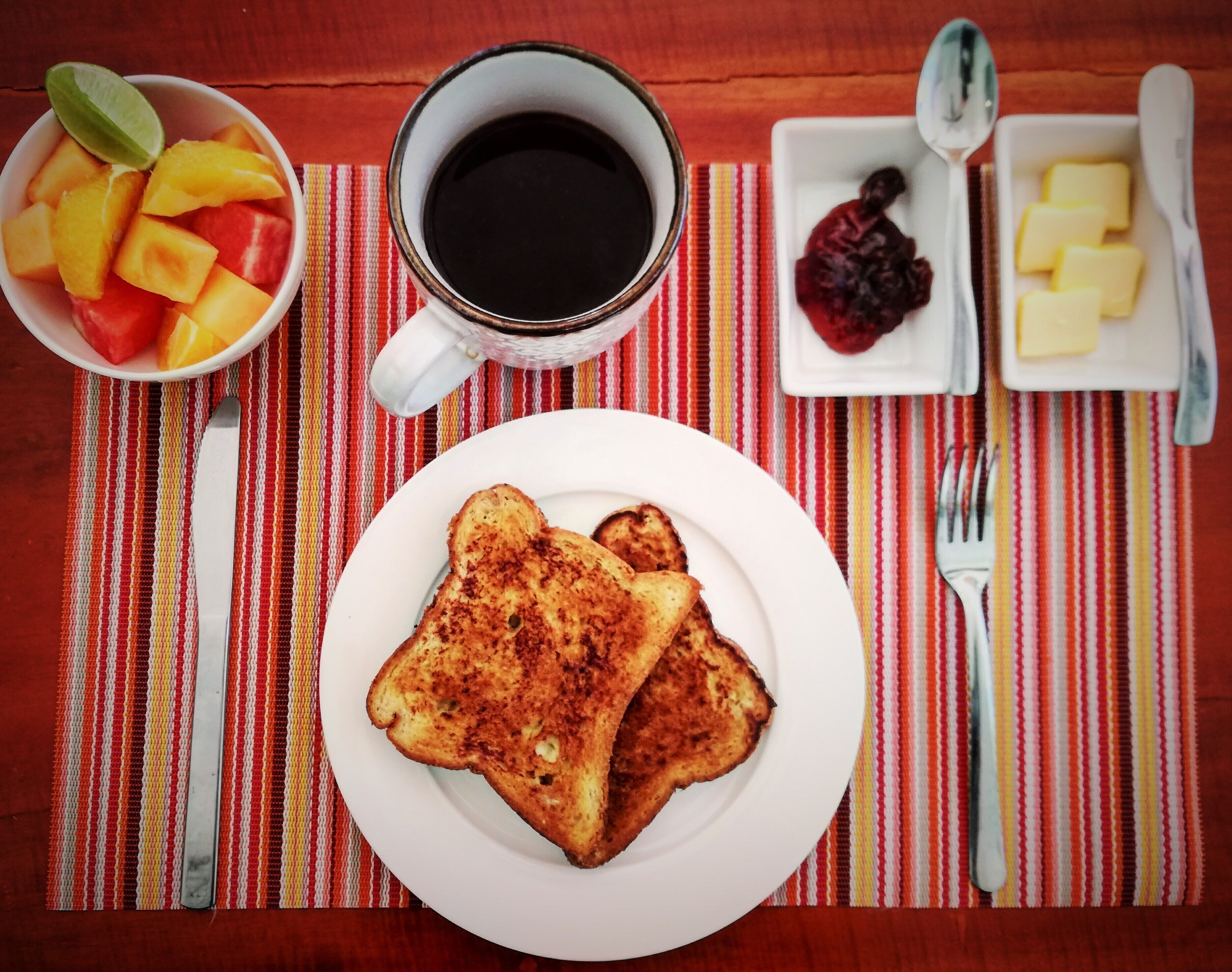 Desayuno con tostadas, una taza de café, un bol de fruta y mantequilla sobre un mantel a rayas.
