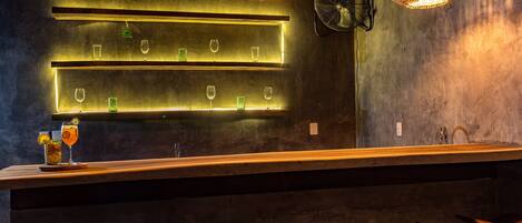 Una zona de bar con una barra larga, varios estantes con vasos y tres lámparas colgantes de mimbre.