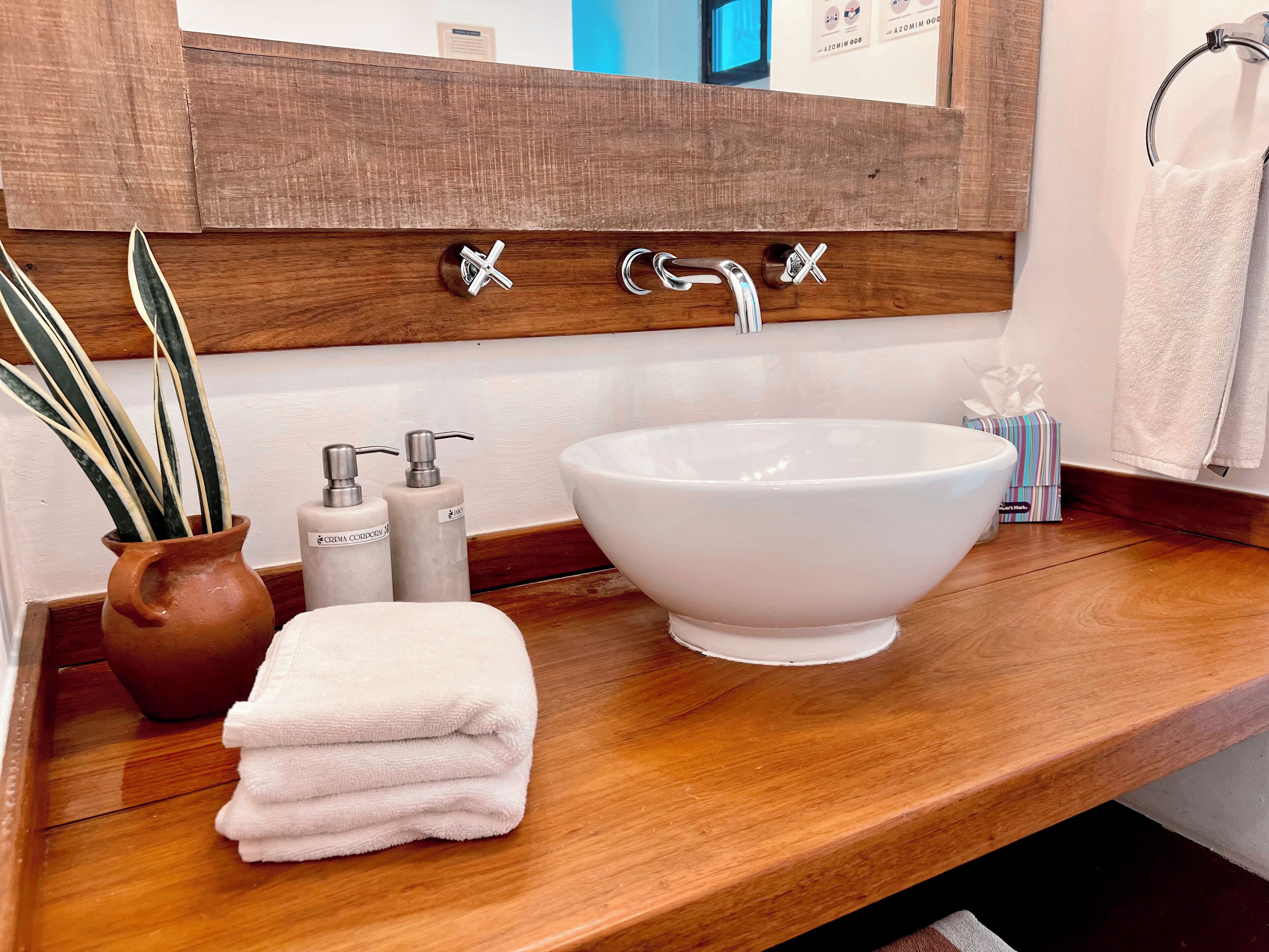A bathroom with a wooden vanity, a white sink, a mirror, a potted plant, and toiletries.