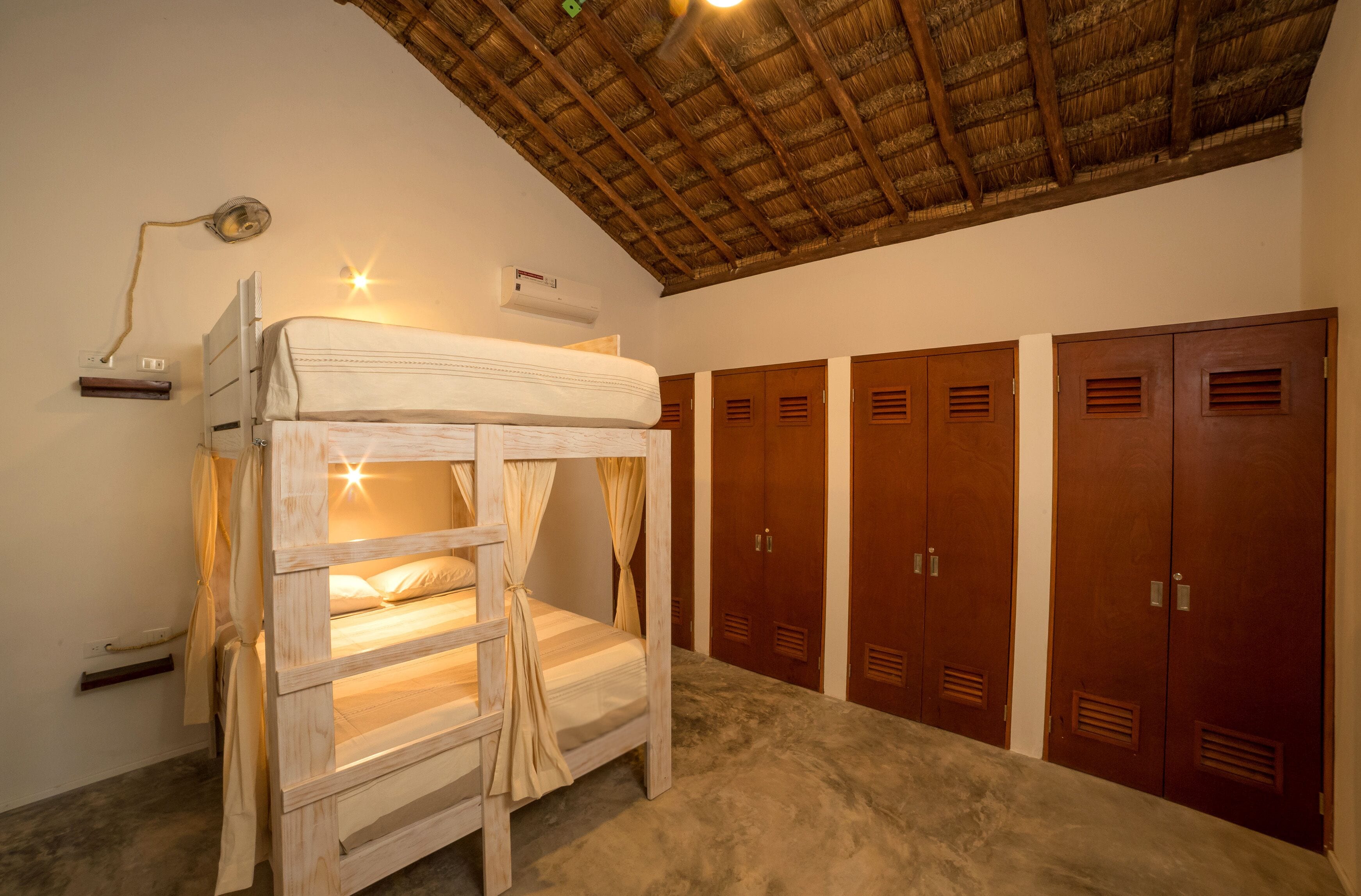 A bunk bed with a ceiling fan, a desk lamp, and a wall-mounted heater. There are wooden lockers and a concrete floor.