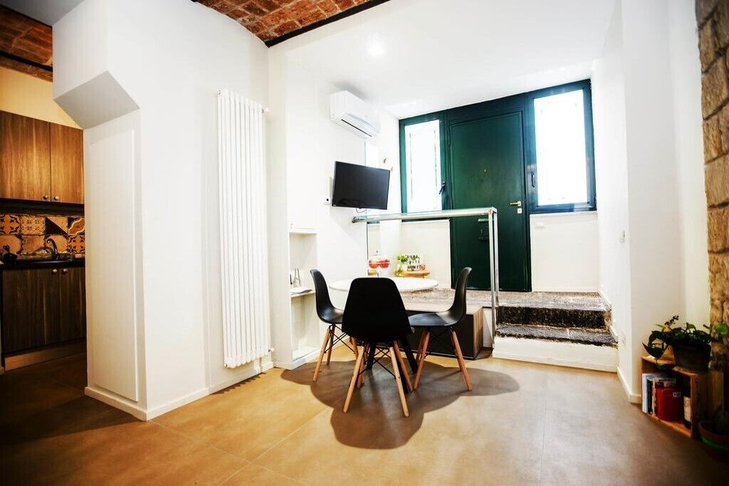 Un ambiente compatto con zona pranzo, televisione a parete, una porta verde e una scala che conduce a un livello superiore.