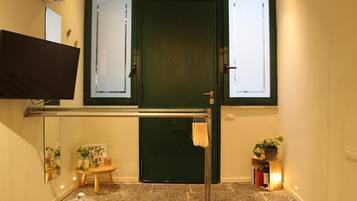 Une pièce avec une porte verte, un téléviseur à écran plat fixé au mur, une petite table en bois sur laquelle est posée une pancarte, une plante en pot et une étagère remplie de livres et de bouteilles.