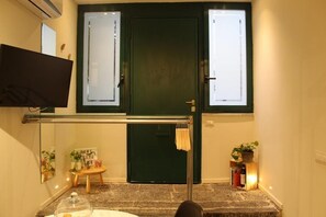Una stanza con una porta verde, una televisione a schermo piatto montata a parete, un piccolo tavolo in legno con un cartello, una pianta in vaso e uno scaffale con libri e bottiglie.