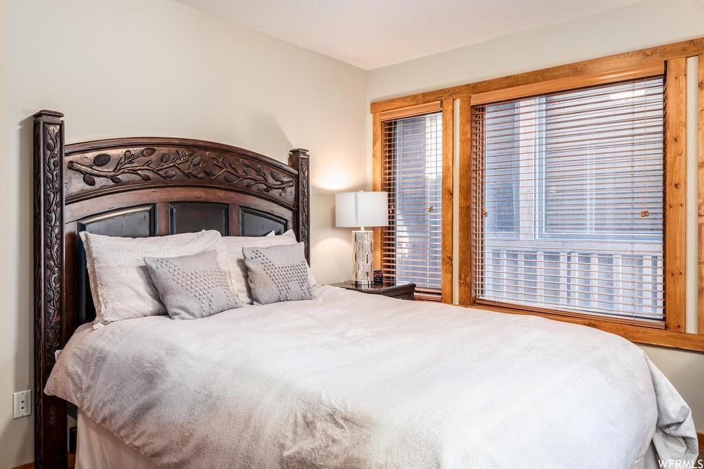 A bedroom with a wooden headboard, a large bed with white bedding, a bedside table with a lamp, and a window with blinds.