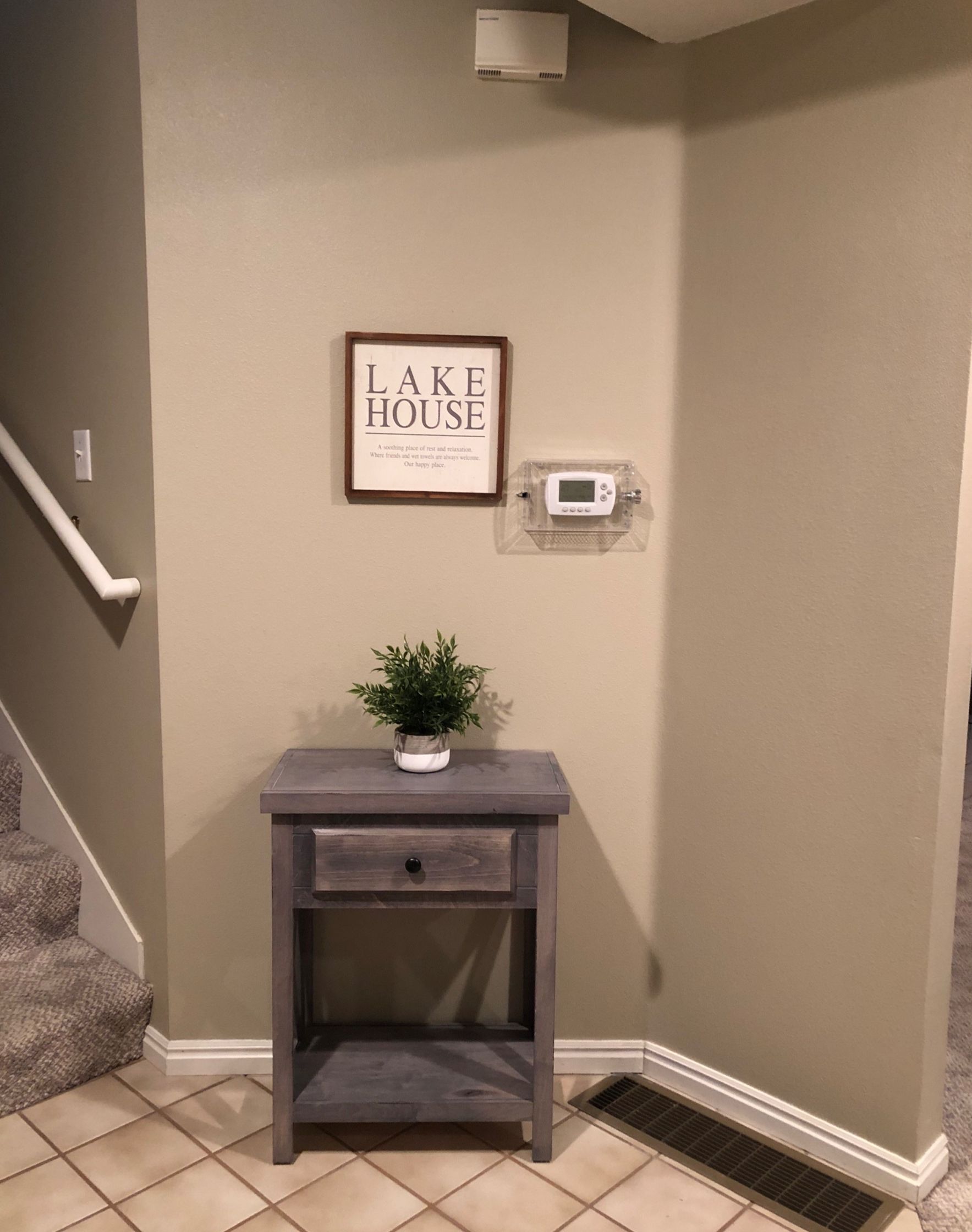 Un pasillo con un letrero enmarcado que dice 'LAKE HOUSE', una pequeña mesa con una planta y un termostato en la pared.