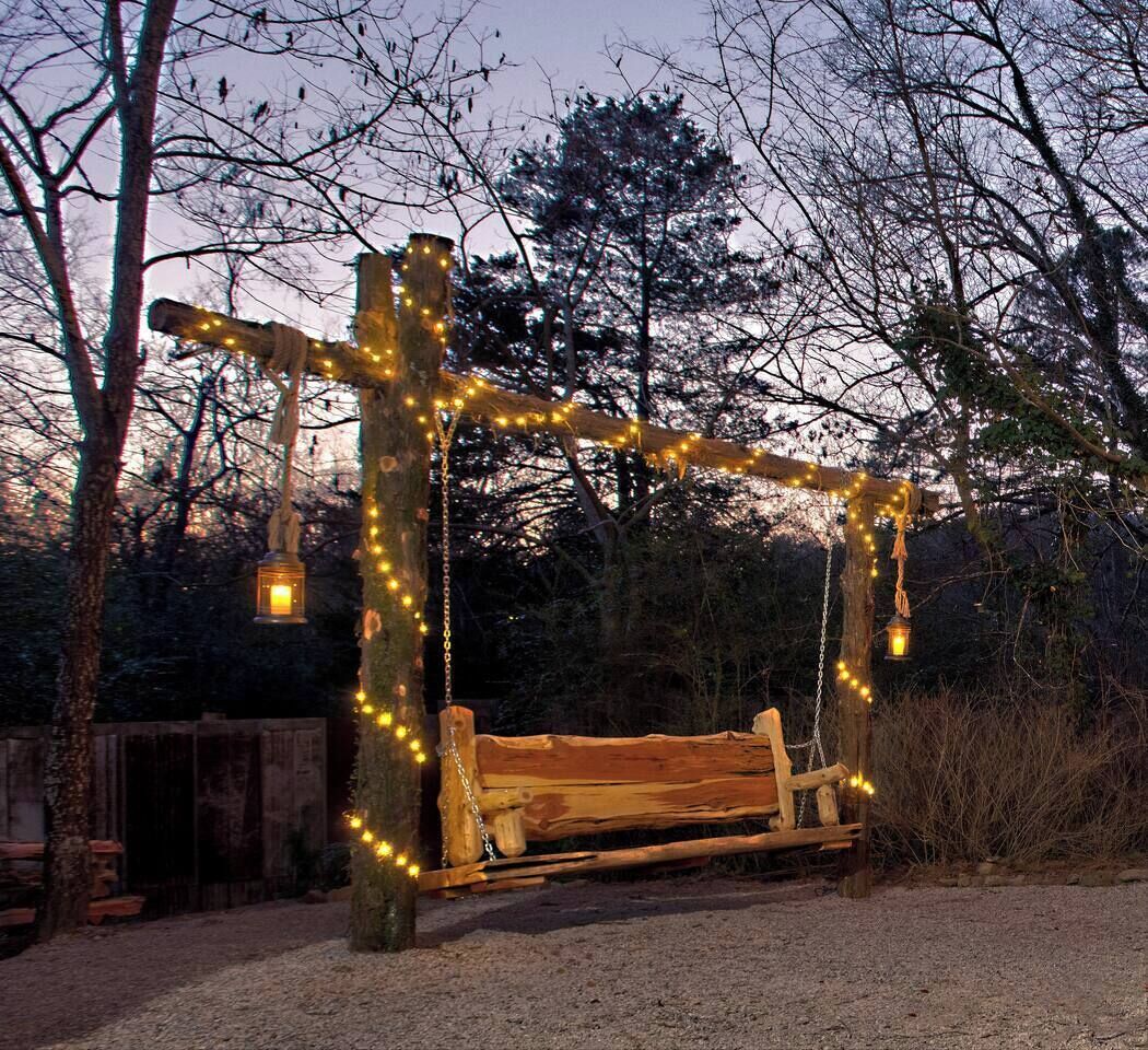 Een houten schommel met verlichting, omgeven door bomen en een grindpad.