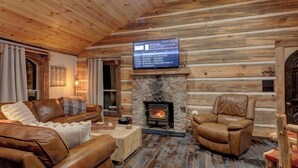 L’intérieur d’un chalet confortable, avec des murs en rondins de bois, une cheminée en pierre, des meubles en cuir et un téléviseur fixé au mur.