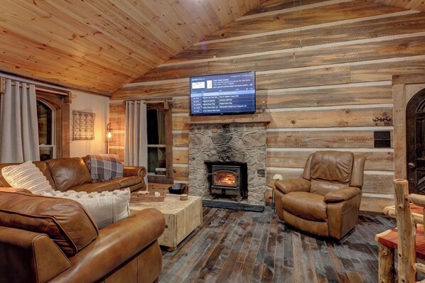 A cozy log cabin interior with a stone fireplace, leather furniture, and a mounted television.