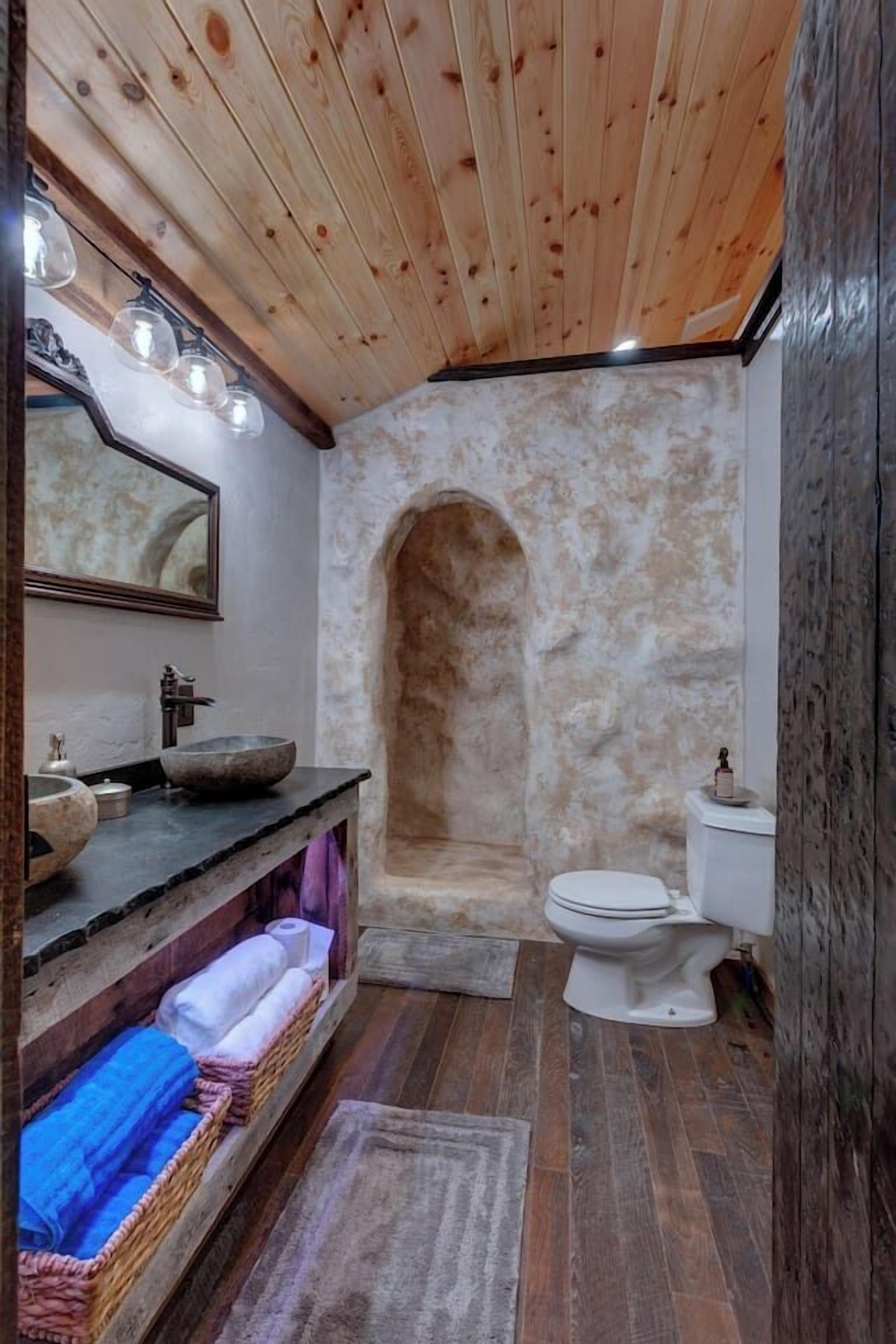 A rustic bathroom with a stone wall, wooden ceiling, and a wooden vanity with a bowl sink.
