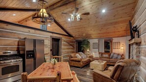 L’intérieur chaleureux d’un chalet en bois comprend un coin repas, deux canapés en cuir, une table à manger en bois, un réfrigérateur, une cuisinière et un ventilateur de plafond.