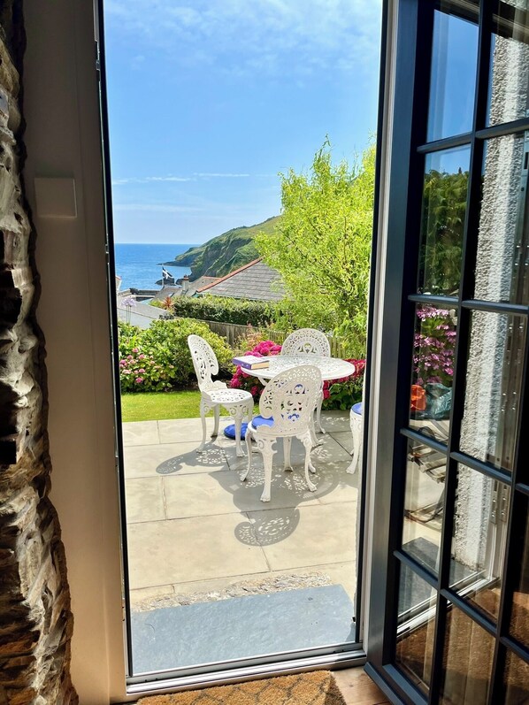 A patio with white chairs and a table, a stone wall, and a view of the sea and hills.