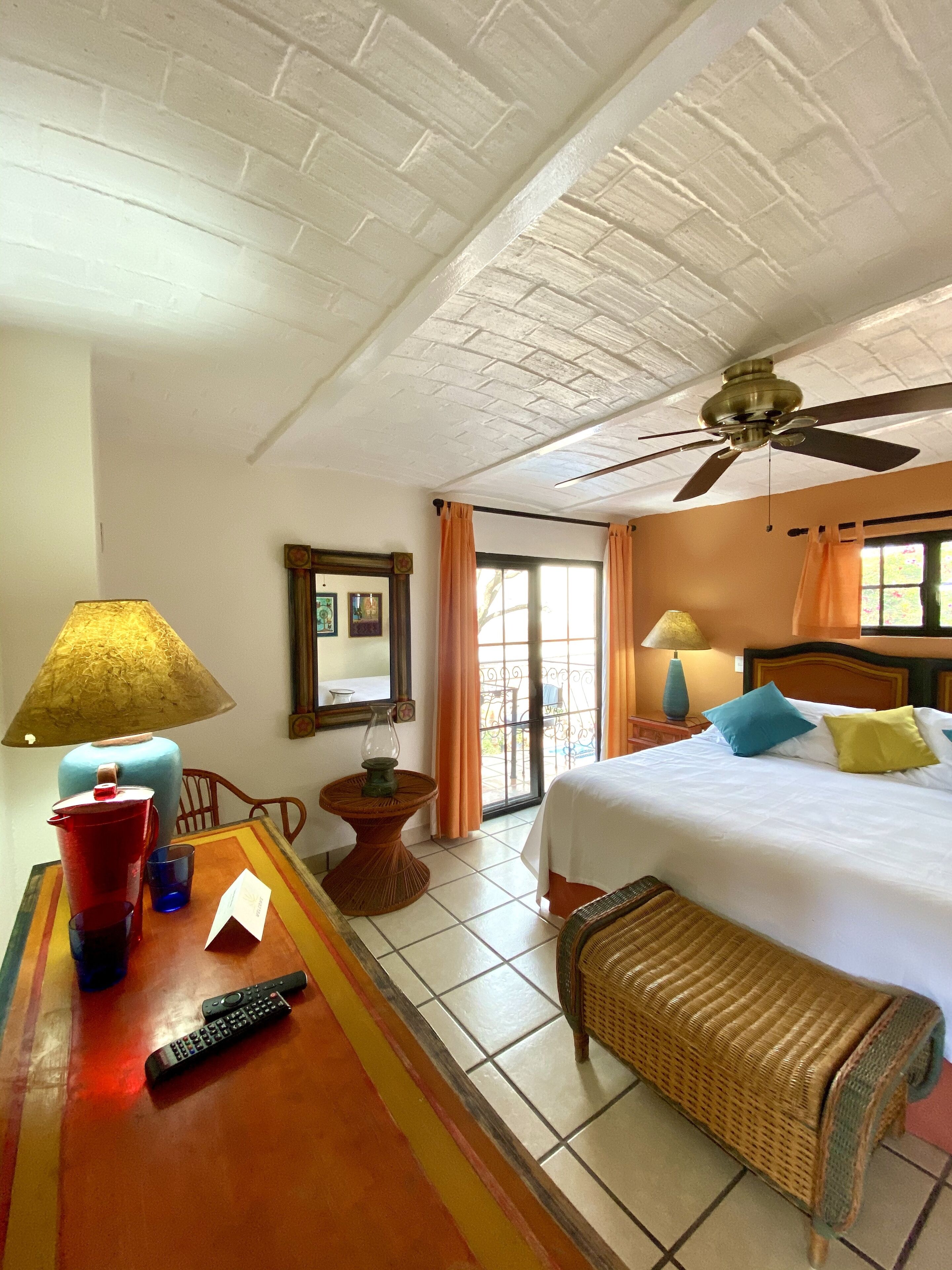 A hotel room with a bed, two lamps, a table with a red pitcher, a remote control, and a wicker bench.