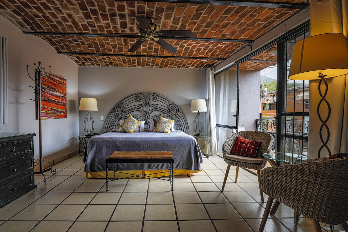 A bedroom with a brick ceiling, a large bed with a headboard, two bedside lamps, a bench, a wicker chair, and a glass table.