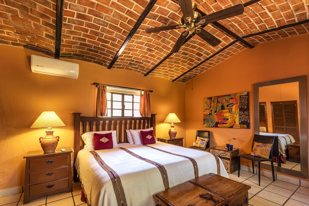 A hotel room with a brick ceiling, a large bed with a wooden headboard, two bedside tables with lamps, a wooden chest, and a mirror on the wall.