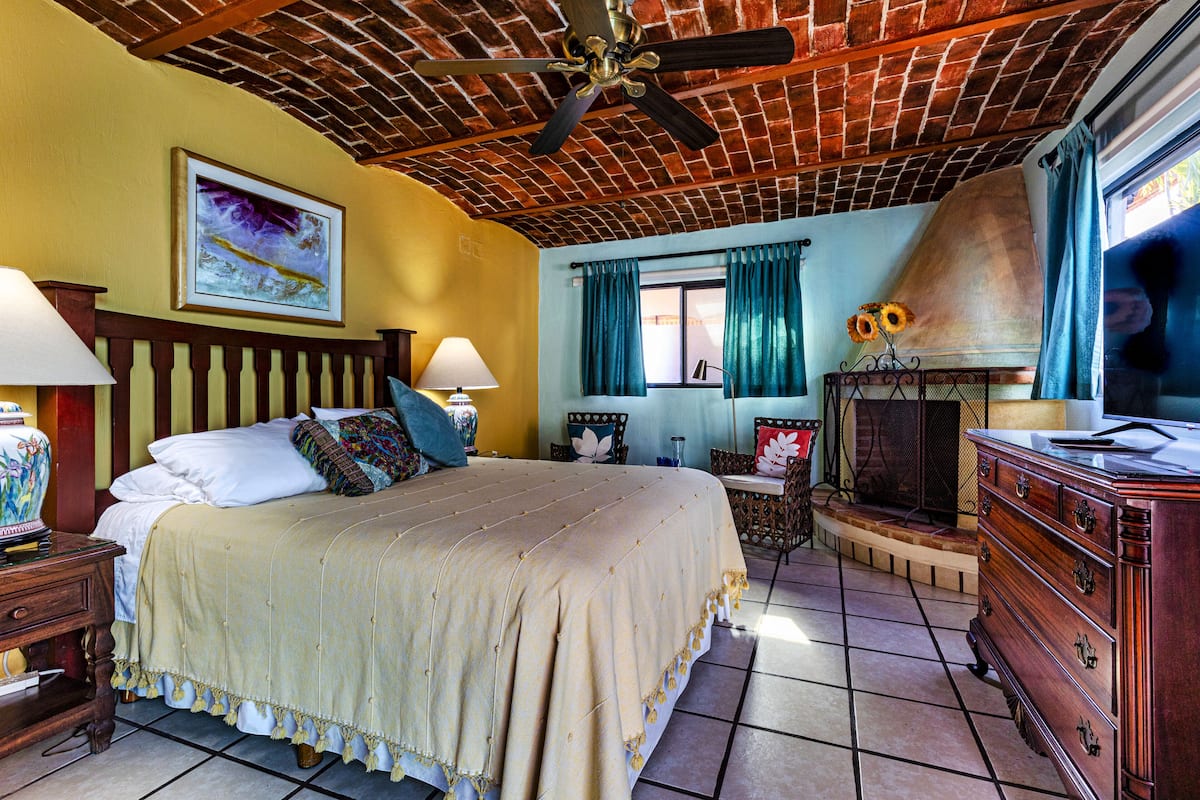 A well-furnished bedroom with a large bed, a wooden dresser, a ceiling fan, and a window with curtains.