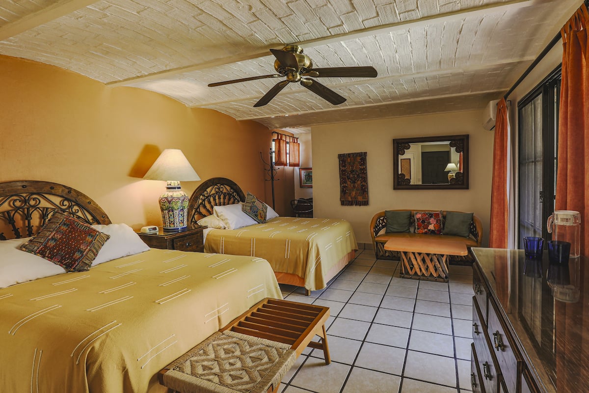 A hotel room with two beds, a ceiling fan, a small table, and a window with curtains.