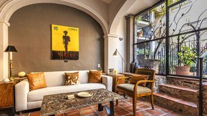 Un salon accueillant avec un canapé blanc, une table basse à motifs et une chaise en bois. Une grande fenêtre donne sur des plantes et un balcon.