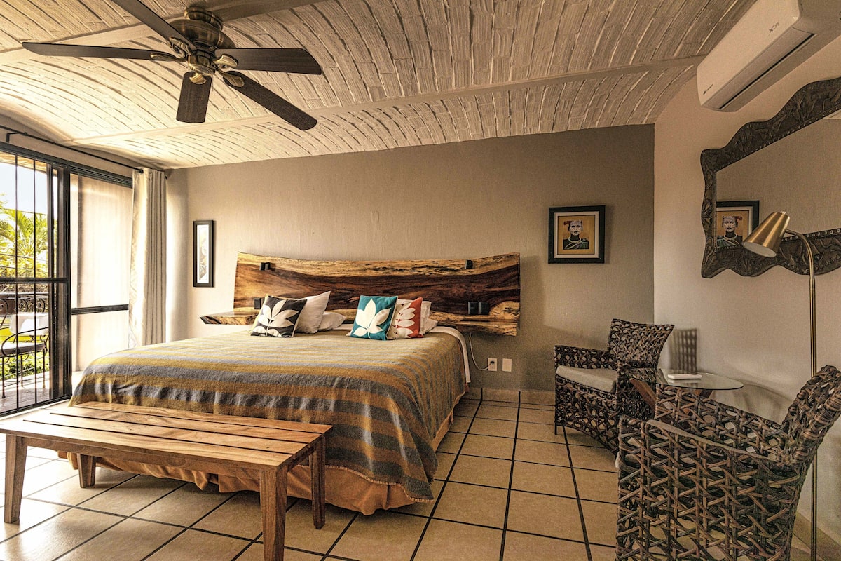A bedroom with a bed, a wooden bench, a chair, a ceiling fan, and a large window.