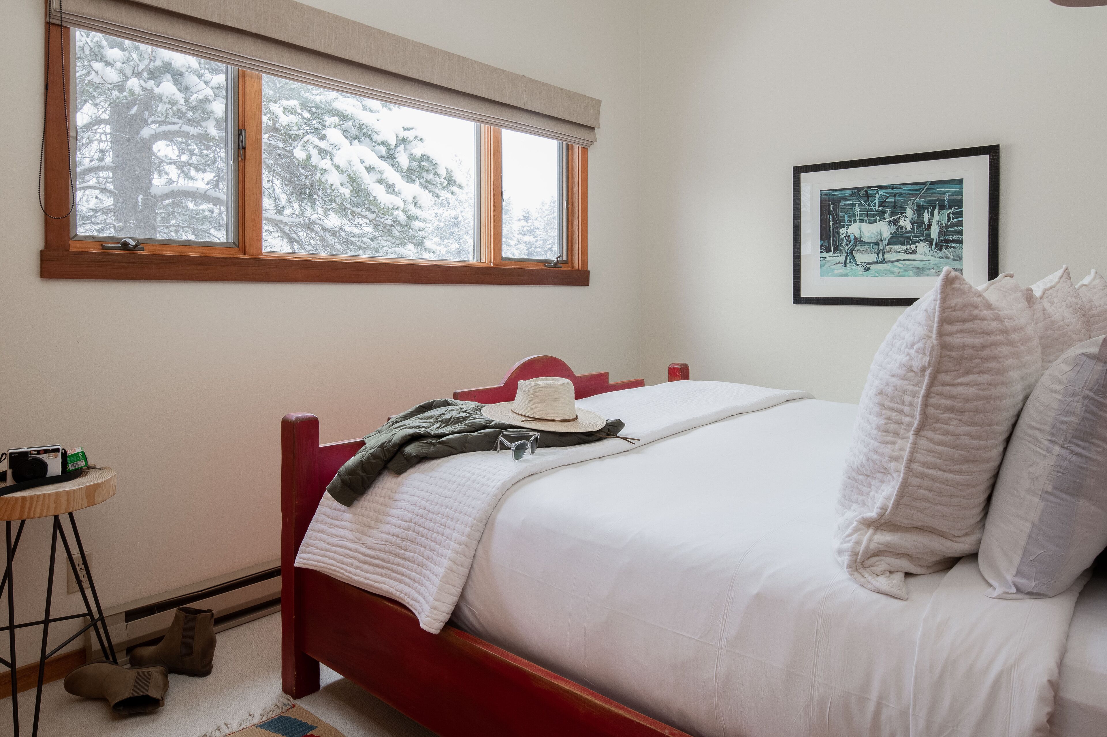 A neatly made bed with a white comforter and pillows, a hat, and a jacket on the side. A framed picture hangs on the wall, and a window with a view of snow-covered trees is visible.