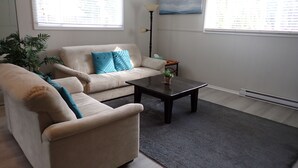 A living room with a beige sofa, a coffee table, and a floor lamp. There are two windows with blinds, a painting on the wall, and a potted plant. - Spacious 2 bedroom suite on private lot (Pemberton)