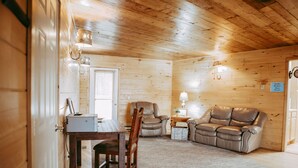 A cozy cabin interior with wooden walls, a dining area with a table and chairs, a leather sofa, and a small side table with a lamp. - Experience Starbuck charm in our spacious 5-bedroom retreat (Starbuck)