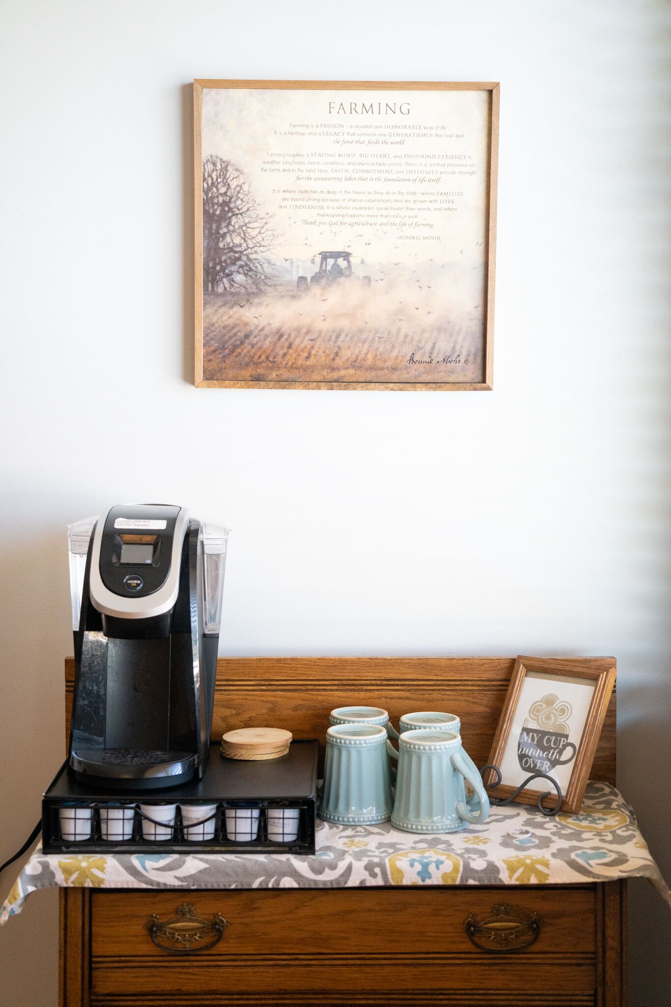 A coffee station with a coffee maker, mugs, and a framed sign on a wall.