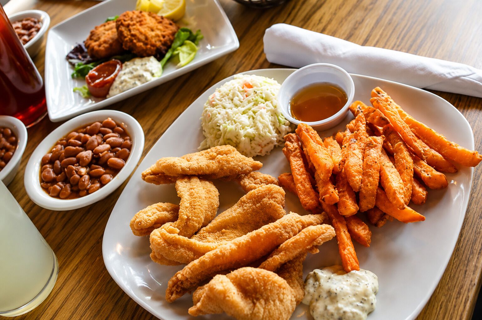 Un plato de pescado frito, papas dulces fritas, ensalada de col y frijoles sobre una mesa de madera.