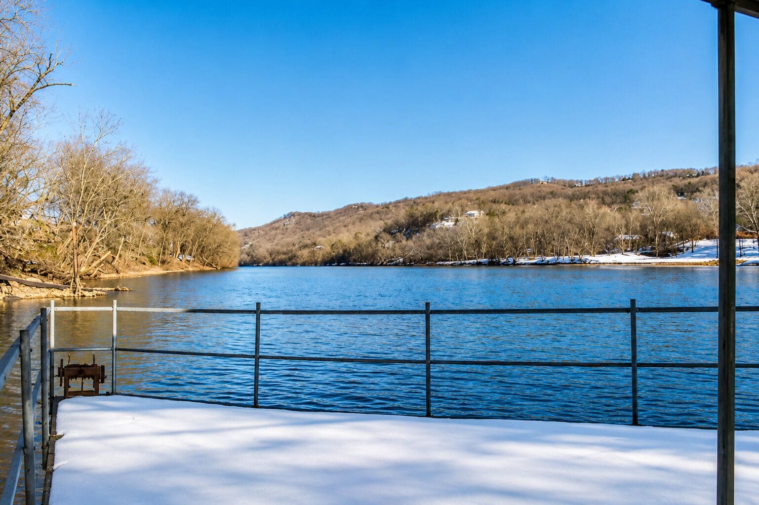 A snow-covered deck with a railing overlooking a river, trees, and a hilly landscape.